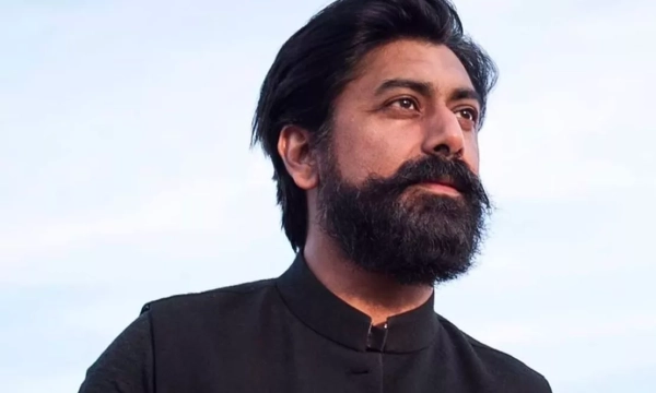 A man with a thick beard and mustache, wearing a black traditional outfit, gazes thoughtfully into the distance against a cloudy sky backdrop.