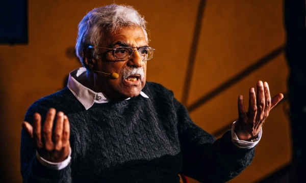 Elderly man with gray hair and glasses passionately speaking, wearing a dark sweater, gesturing with hands in a conversation setting.