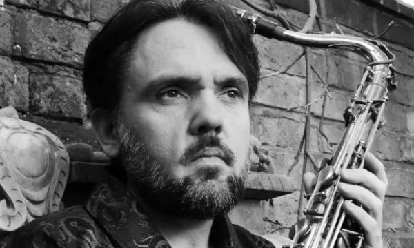 A solemn man with a beard holds a saxophone, gazing pensively off to the side, set against a textured brick wall.