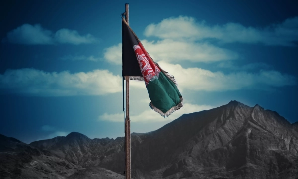 Afghan flag fluttering on a flagpole with a mountainous landscape and blue sky in the background.