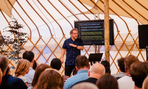 A speaker in a tent setting discusses audience reactions, with a screen displaying key questions for engagement. Attendees are seated, intently listening.