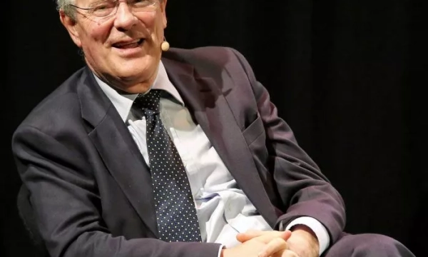 A middle-aged man in a suit smiles while seated on stage, with a microphone headset, against a dark backdrop.