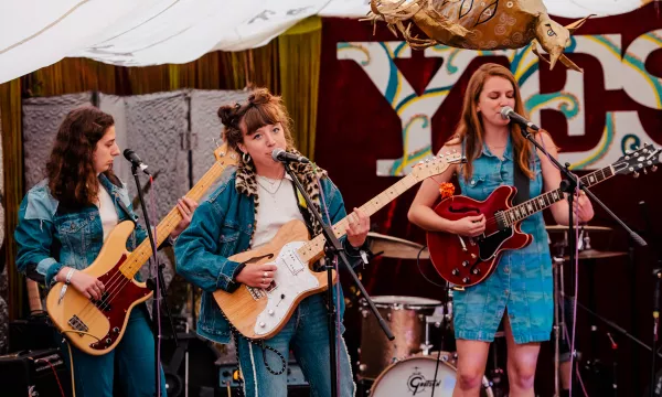 A live music performance featuring three female musicians in denim outfits, with one playing guitar and the others on bass and vocals, set in a decorated venue.