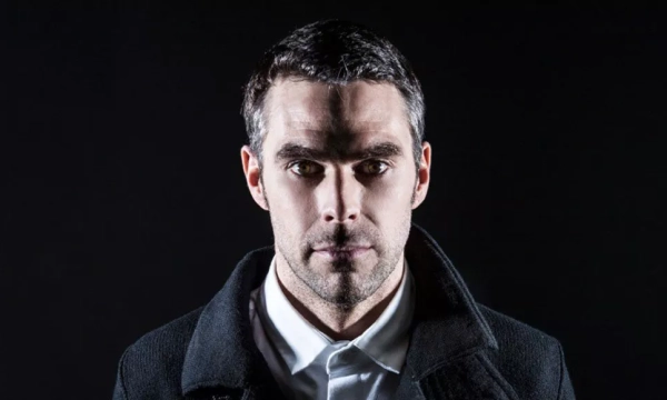 A serious-looking man with short dark hair, wearing a black coat and white shirt, is illuminated against a dark background.