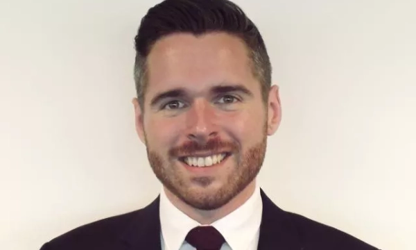 A well-dressed man with short, styled hair and a beard smiles at the camera, wearing a suit and tie against a plain background.