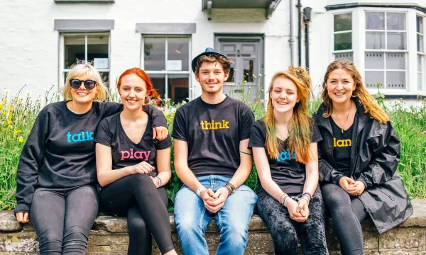 A group of five young adults in casual black t-shirts with colorful words ("talk," "think," "play," "wild," "love") sit on a stone wall in front of a white building.
