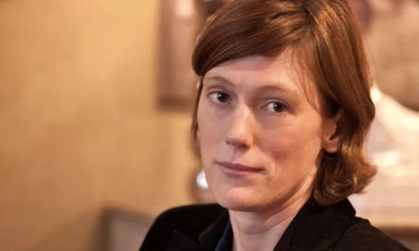 A woman with shoulder-length brown hair and a serious expression is seated, wearing a black jacket against a softly blurred background.