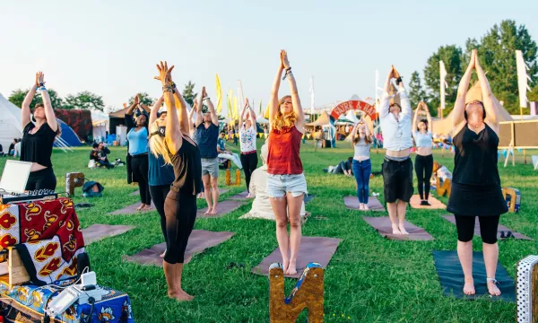 A diverse group of people practicing yoga outdoors, raising their arms in a sun salutation on yoga mats in a grassy field during a festival.