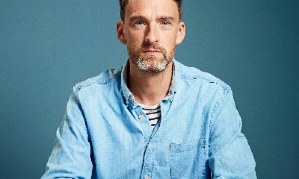 A man with short, curly hair and a beard wears a light blue shirt, seated at a table against a solid blue background.