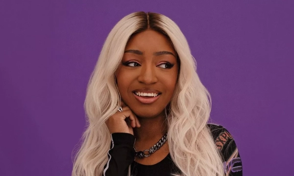 A smiling person with long, wavy blonde hair dressed in a stylish outfit, posed against a vibrant purple background.