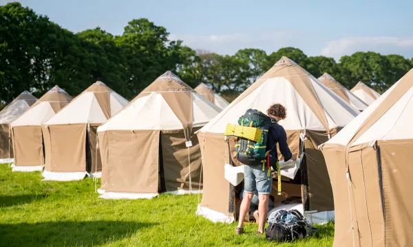 A person with a backpack is opening a glamping tent in a green field, with several similar tents in the background.