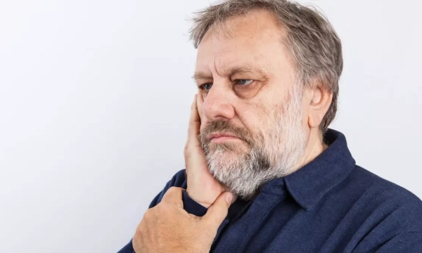 A contemplative man with gray hair and a beard touches his face, wearing a dark blue shirt against a plain white background.